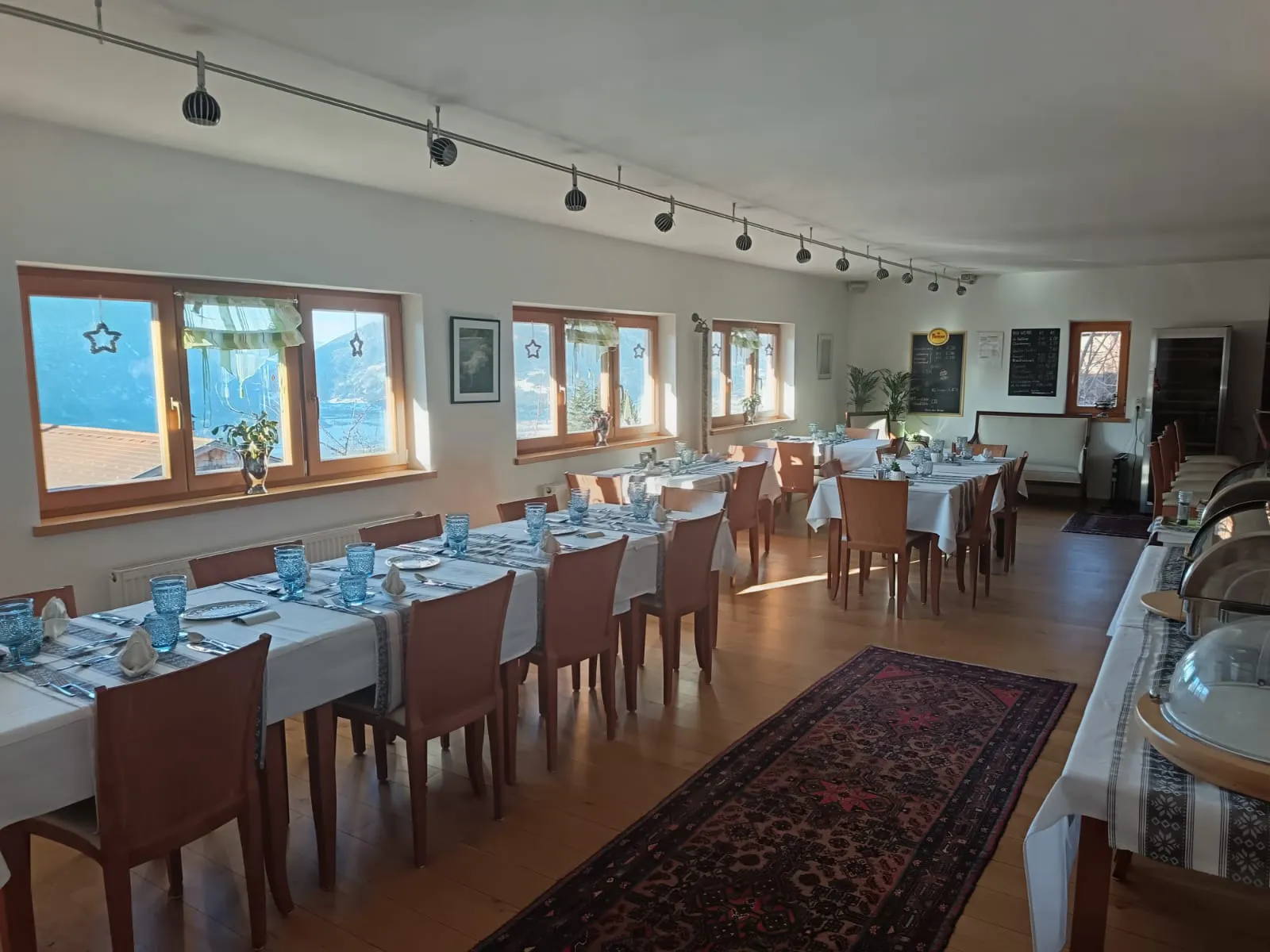 Geräumiger Speisesaal mit mehreren gedeckten Tischen, mit weißen Tischdecken, blauen Trinkgläsern und ordentlich angeordnetem Besteck. Der Raum verfügt über große Fenster, die Tageslicht hereinlassen, mit sternförmigen Dekorationen, Holzstühlen und einem gemusterten Teppich auf dem Holzboden. Die Buffetgerichte sind abgedeckt und stehen auf einem Beistelltisch.